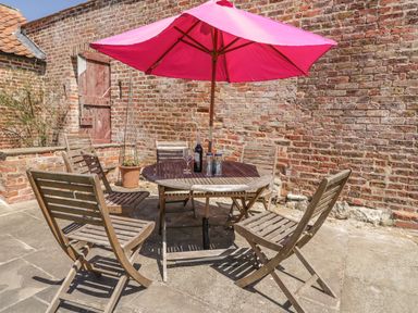 An outdoor patio with a wooden table and chairs under a pink umbrella with a brick wall background at Cow Byre Malton