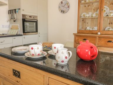 A kitchen countertop with cups saucers a red teapot biscuits a wooden cabinet with dishes and a wall clock at Cow Byre Malton