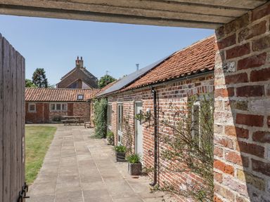 A brick building with a red tiled roof and paved pathway with plants and a wooden table at Cow Byre in Malton