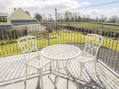A balcony with a white metal table and two chairs overlooking a garden and a house at Oak Cottage in Bryngwenith near Newcastle Emlyn