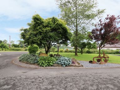 A garden island with trees plants and a rusty old farming plow on a gravel area by a paved road at Oak Cottage in Bryngwenith near Newcastle Emlyn