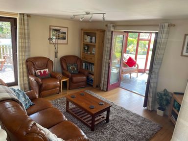A living room with leather armchairs a wooden coffee table a bookshelf and a door leading to a sunroom with wicker furniture at Oak Cottage in Bryngwenith near Newcastle Emlyn
