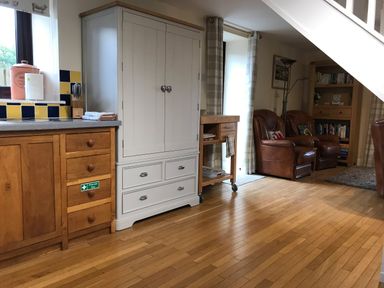 A kitchen area with wooden cabinets and a countertop next to a white cupboard and two brown leather armchairs with a wooden bookshelf behind them at Oak Cottage in Bryngwenith near Newcastle Emlyn
