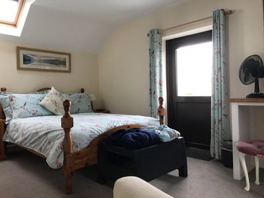 A bedroom with a wooden bed covered with floral bedding a black ottoman with clothes a window and curtains a desk with a fan and a pink stool at Oak Cottage in Bryngwenith near Newcastle Emlyn