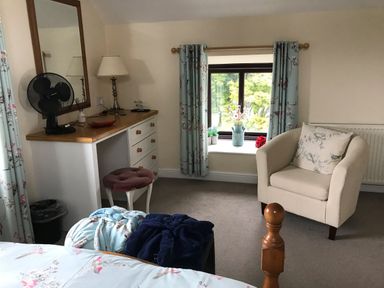 A bedroom corner with a dressing table fan lamp and stool beside a window with floral curtains and a beige armchair at Oak Cottage in Bryngwenith near Newcastle Emlyn