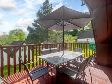 An outdoor wooden deck with a table and six chairs under a large umbrella surrounded by trees and houses at Brookside Honicombe Manor near Gunnislake