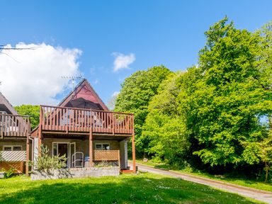 A wooden cabin with a balcony and outdoor furniture surrounded by green trees at Brookside in Honicombe Manor near Gunnislake
