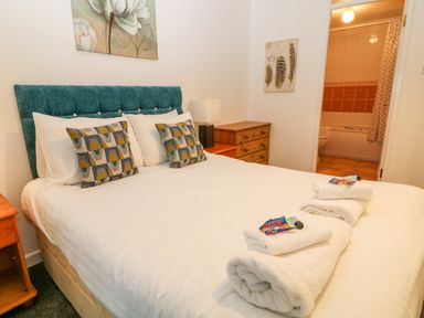A bedroom with a double bed with geometric pillows white towels on the bed a wooden dresser a nightstand with a lamp and an open door to a bathroom at Brookside in Honicombe Manor near Gunnislake