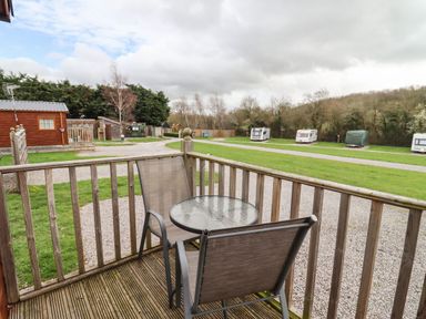 A deck with two chairs and a round glass table overlooking a grassy area with caravans and a wooden cabin at Broad Down in Berrow