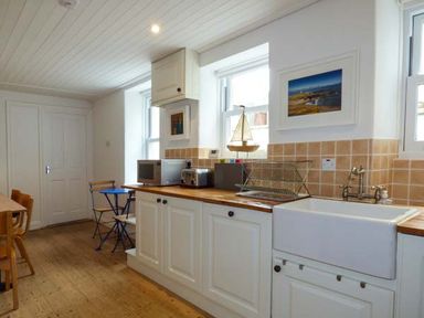A kitchen with wooden countertops white cabinets a white sink a drying rack a microwave a toaster two windows and a small blue table with chairs at Sandy Bank in Rhosneigr