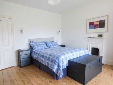 A bedroom with a double bed with blue plaid bedding two bedside tables a wooden floor and a fireplace at Sandy Bank in Rhosneigr