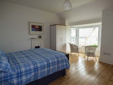 A bedroom with a blue plaid bedspread a white wardrobe two chairs and a window at Sandy Bank in Rhosneigr