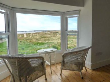 A sitting area with two wicker chairs and a small table by large windows overlooking grass and sandy dunes at Sandy Bank in Rhosneigr