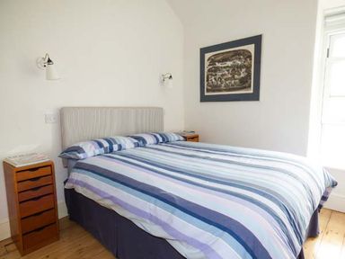 A bedroom with a bed with striped blue bedding a wooden drawer unit wall lights and a framed picture on the wall at Sandy Bank in Rhosneigr