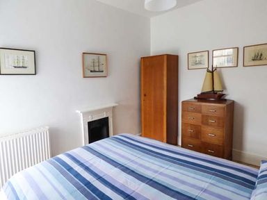 A bedroom with a striped blue bedspread a wooden wardrobe a chest of drawers with a model ship and ship pictures on the wall at Sandy Bank in Rhosneigr