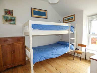 A bedroom with a white bunk bed with blue bedding a wooden cabinet a chair and paintings on the wall at Sandy Bank in Rhosneigr