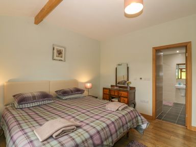 A bedroom with a large bed covered in plaid bedding a wooden floor a wooden dressing table with a mirror a lamp and an open door leading to a bathroom at Caer Moel in Caernarfon