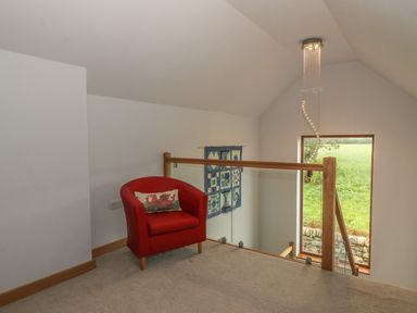 A small seating area with a red armchair and a cushion next to a wooden and glass railing overlooking a stairwell with a window showing greenery at Caer Moel in Caernarfon