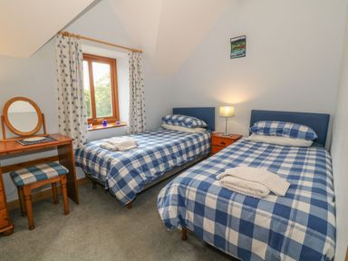 A bedroom with two single beds with blue checkered bedding a wooden vanity with a round mirror and stool and a window with floral curtains at Caer Moel in Caernarfon