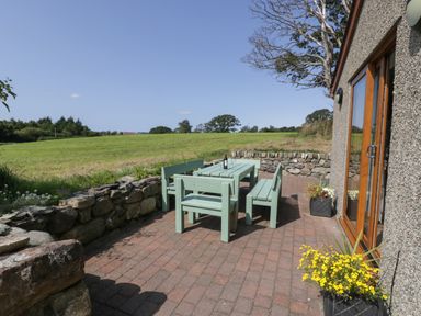 A patio with a green wooden table and benches on a brick surface next to a stone wall and a grassy field at Caer Moel in Caernarfon