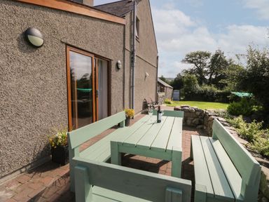 An outdoor patio with green wooden benches and a table with a bottle and glass next to a stone wall at Caer Moel in Caernarfon