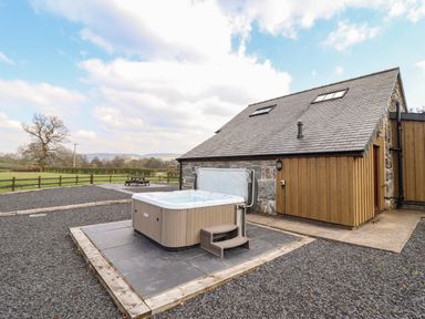 An outdoor hot tub on a tiled platform next to a stone and wood building with a fenced field in the background at Beudy Bach Barn in Llanuwchllyn near Bala