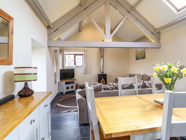A dining table with chairs and flowers in a room with exposed beams a wood stove a sofa and a television at Beudy Bach Barn in Llanuwchllyn near Bala