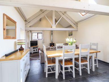 A dining area with a wooden table and six chairs facing a living room with a sofa television and a wood stove at Beudy Bach Barn in Llanuwchllyn near Bala