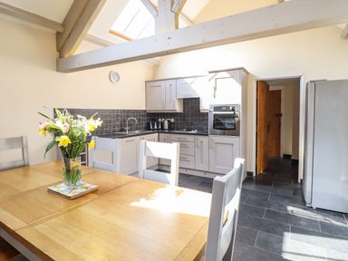 A kitchen with a wooden dining table and chairs a vase with flowers on the table white cabinets and a built-in oven a refrigerator and a tiled floor at Beudy Bach Barn in Llanuwchllyn near Bala