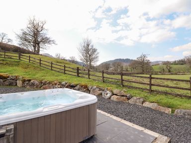 An outdoor hot tub next to a gravel path with a wooden fence and grassy hills in the background at Beudy Bach Barn in Llanuwchllyn near Bala
