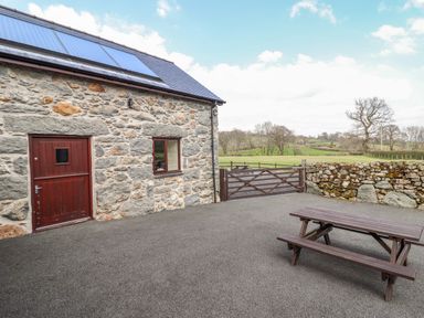 A stone building with a wooden door and window a wooden picnic table a stone wall and gate with green fields beyond at Beudy Bach Barn in Llanuwchllyn near Bala