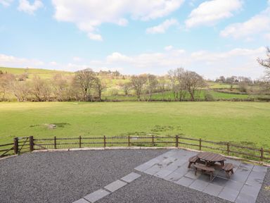 A fenced outdoor area with gravel and tiled patio with round wooden picnic table overlooking green fields and trees at Beudy Bach Barn in Llanuwchllyn near Bala