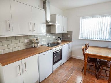 A kitchen with white cabinets wooden countertops a stove with an oven a kettle a small fridge a wooden dining table with four chairs and a window with vertical blinds at Arfryn in Bethesda