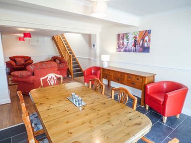 A dining room with wooden table and chairs next to a sideboard and red armchairs with a staircase and red sofas in the background at Arfryn in Bethesda