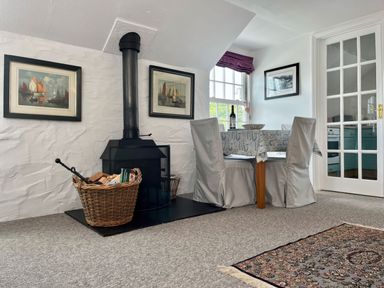 A dining area with a small table and two covered chairs next to a wood-burning stove and basket with firewood at The Boat House in Golant near Tywardreath