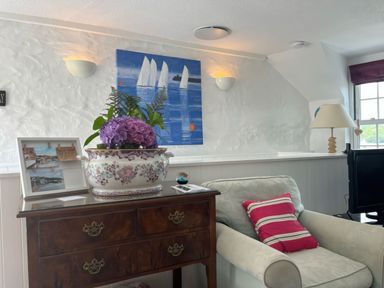 A sitting area with a floral armchair, wooden dresser with a flowerpot and picture frame, and a painting of sailboats on the wall at The Boat House in Golant near Tywardreath
