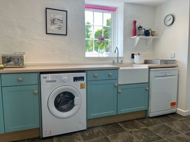 A kitchen with blue cabinets a washing machine dishwasher sink and window with flowers at The Boat House in Golant near Tywardreath