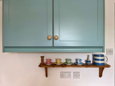 A kitchen wall with a blue cabinet above a wooden shelf holding striped cups saucers and a jug at The Boat House in Golant near Tywardreath