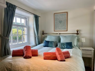 A bedroom with a bed with multiple pillows and red towels on it near a window with curtains at The Boat House in Golant near Tywardreath