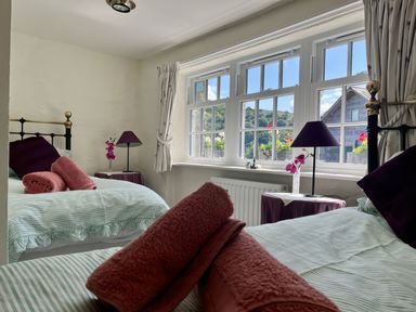 A bedroom with two single beds each with striped bedding and rolled towels with purple lamps and flowers on bedside tables at The Boat House in Golant near Tywardreath
