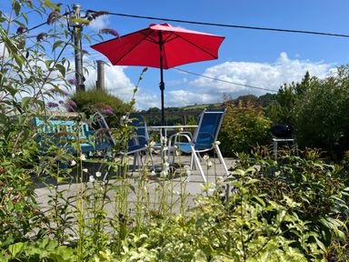 A patio with a red umbrella over a table and blue chairs surrounded by plants and greenery at The Boat House Golant near Tywardreath