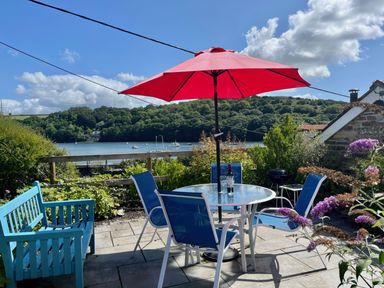 A patio with blue chairs and a table under a red umbrella overlooking a river with boats and green hills at The Boat House in Golant near Tywardreath