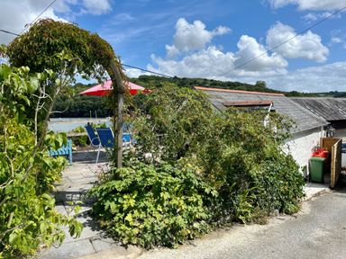A garden patio area with a wooden arch covered in plants blue chairs and a red umbrella near a white building at The Boat House in Golant near Tywardreath