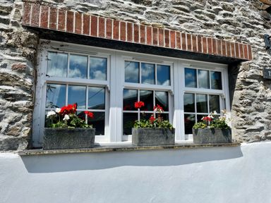 A stone wall with a three-pane window and flower boxes containing red and white flowers at The Boat House in Golant near Tywardreath