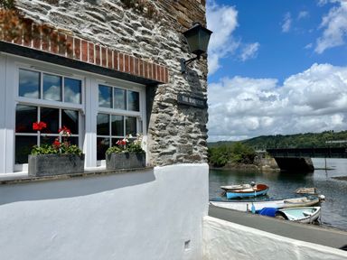 Exterior wall of a stone building with white window frames and flower boxes by a riverside with small boats and a bridge at The Boathouse in Golant near Tywardreath