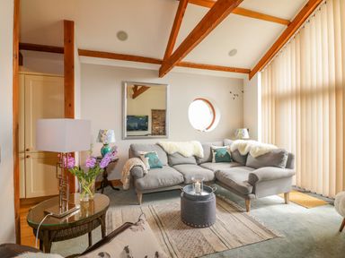 A living room with grey sofas a round window wooden beams and vertical blinds at The Wagon House in Tresmeer near Launceston