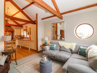 A living room with a gray sectional sofa pillows and a round window next to an open kitchen with wooden beams and white cabinets at The Wagon House in Tresmeer near Launceston