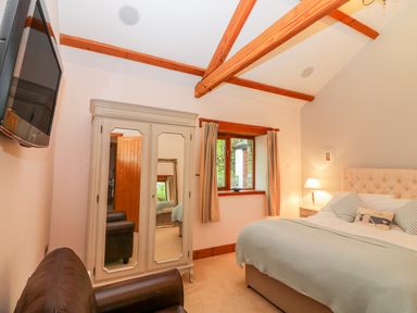 A bedroom with a bed a nightstand lamp a wardrobe with mirrors a window with curtains a mounted television and wooden ceiling beams at The Wagon House in Tresmeer near Launceston