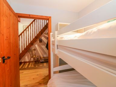 A bedroom with white bunk beds and wooden door opening to a hallway with wooden stairs and chair at The Wagon House in Tresmeer near Launceston