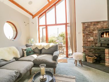 A living room with a gray sectional sofa a round window a lit fireplace and large wooden framed glass doors at The Wagon House in Tresmeer near Launceston